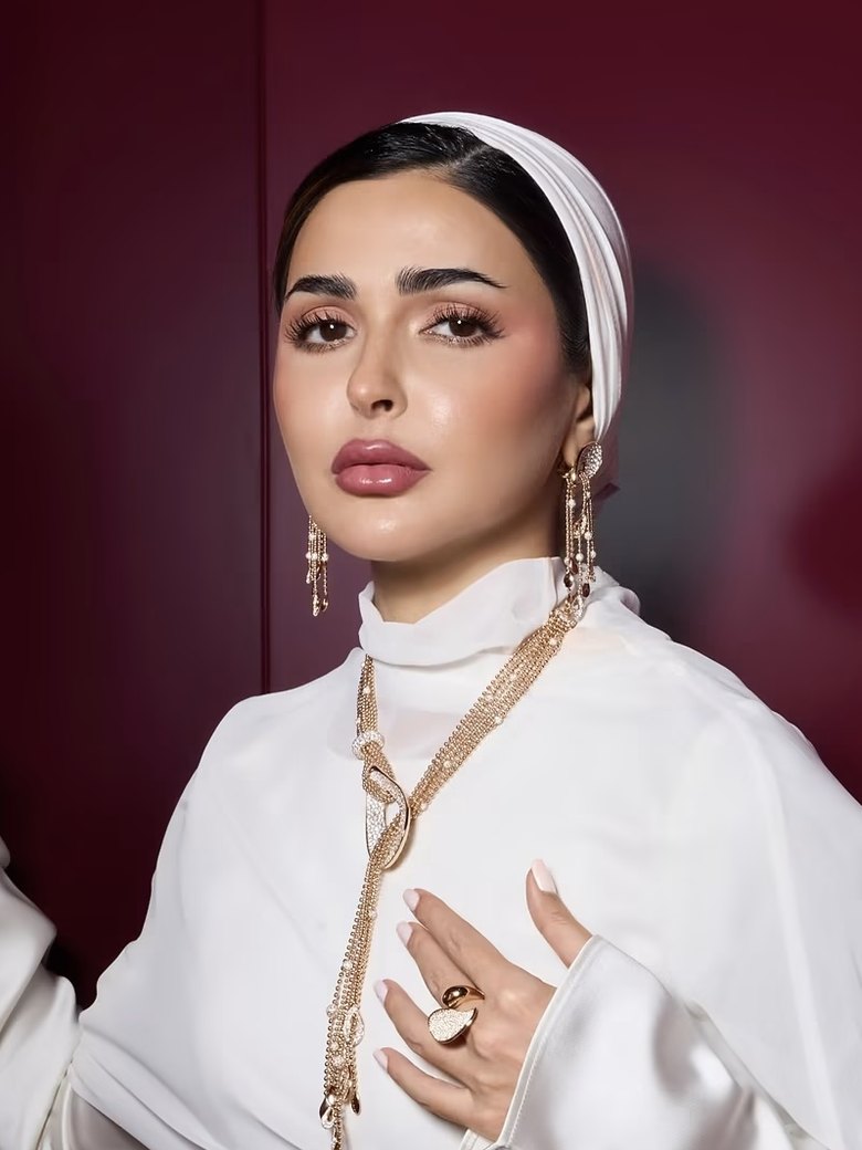 Haneen Al-Saify in a white dress and headwrap against a deep red wall, gold jewellery at her ears and neck