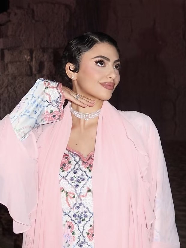 Shayma in a pink abaya at night, her hand raised to her chin, old stone walls behind her
